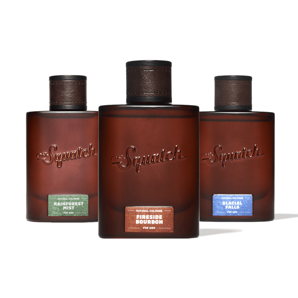 Three Squatch fragrance bottles arranged in a row against a solid green background. The center bottle is upright and labeled 'Squatch Fireside Bourbon', while the left and right bottles are slightly angled backward and labeled 'Squatch Rainforest Mist' and 'Squatch Glacial Falls' respectively.